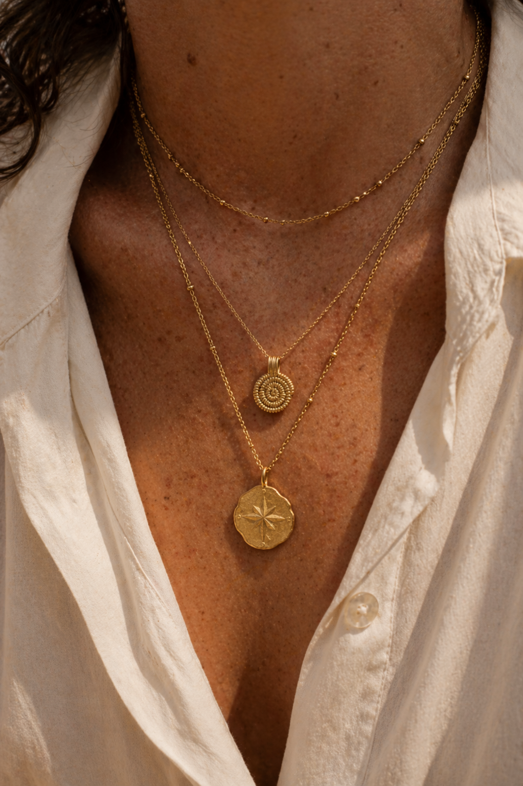 Close-up of a person wearing gold necklaces with circular and star-shaped  compass pendants, against a white shirt background. Made in France 24K gold